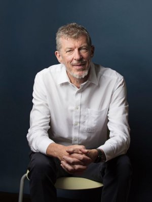 A man with grey hair and wearing a white shirt sits clasping his hands